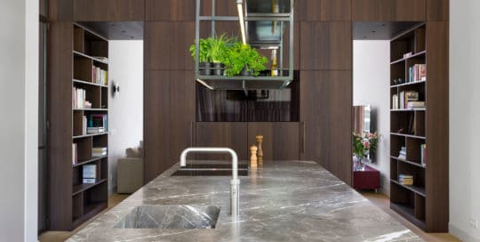 Küchenblock aus FIORE DI BOSCO in Abwicklung mit Küchenrückwand aus Räuchereiche und beleuchtetem Stahlregal über der Küche mit Quooker Wasserhahn